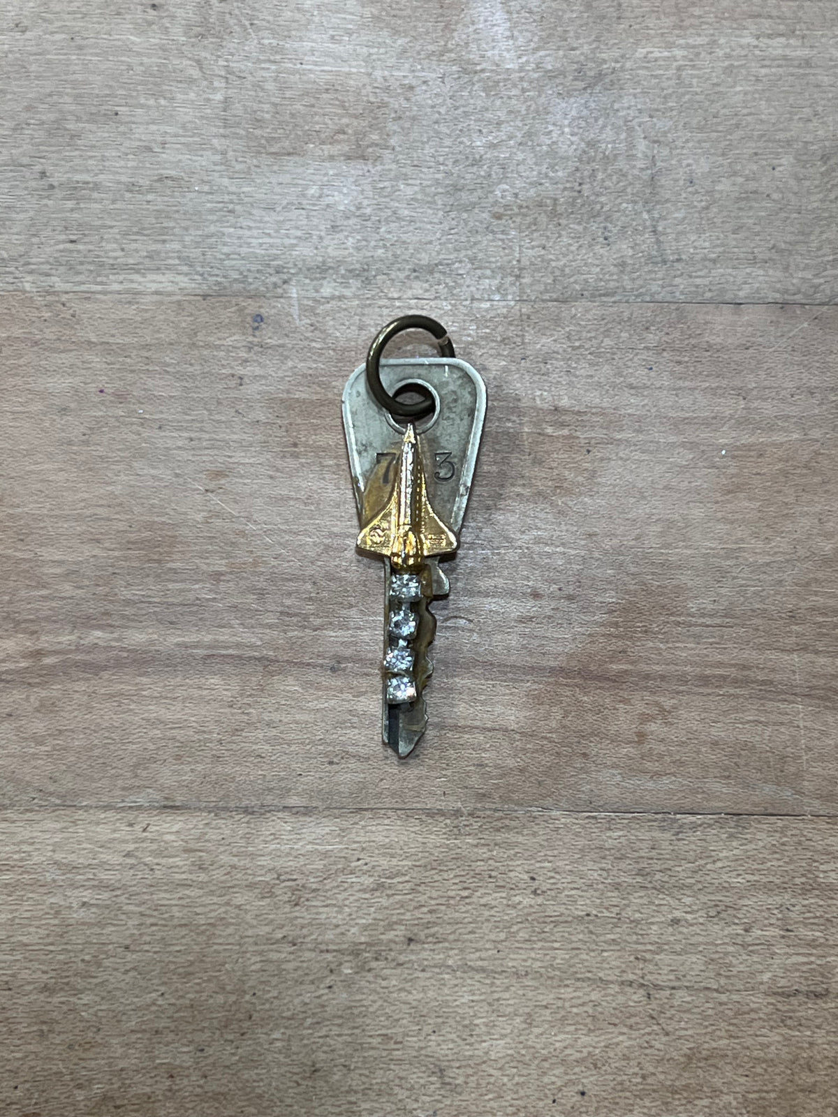 A vintage-style key with a silver-toned metal head and a dangling clear faceted crystal, resting on a light brown wood-grain surface.
