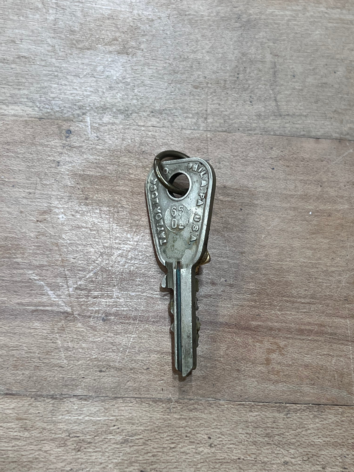 A single vintage metal key with a keyring, photographed on a light brown wood-grain surface.
