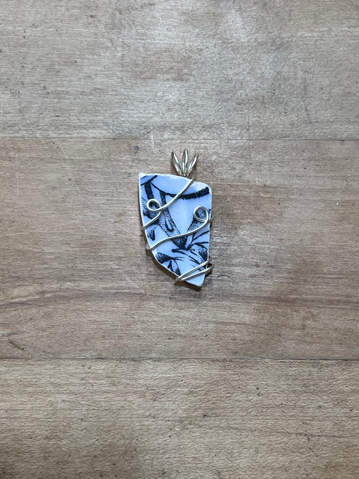 A handmade pendant featuring a light blue and white patterned stone wrapped in silver-colored wire, displayed on a rustic wooden surface.