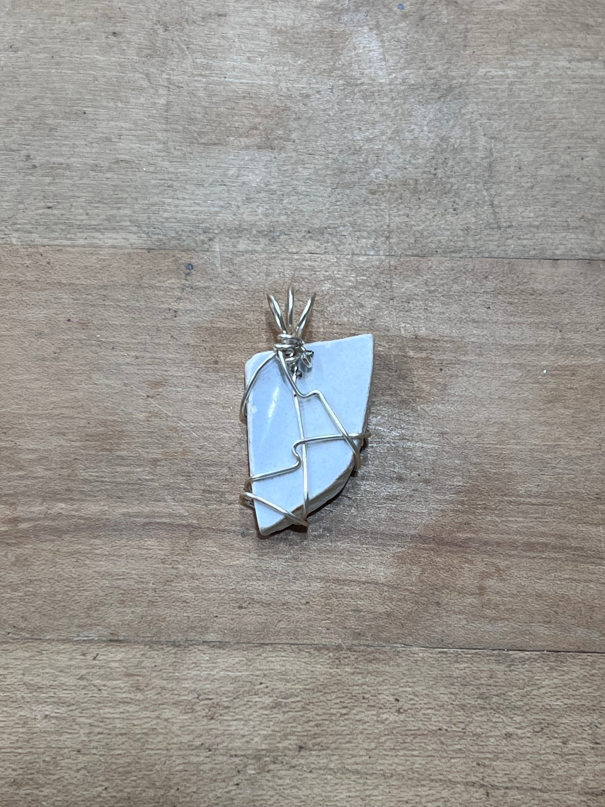 A handmade pendant featuring a light blue-gray irregular stone wrapped in silver-colored wire, resting on a light brown wooden surface.