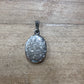 An oval silver pendant with an embossed floral pattern, resting on a light brown wooden surface.