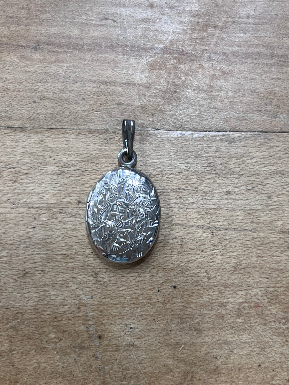 An oval silver pendant with an embossed floral pattern, resting on a light brown wooden surface.