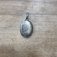 An oval silver pendant with a weathered, patinated surface, resting on a light brown wooden surface.