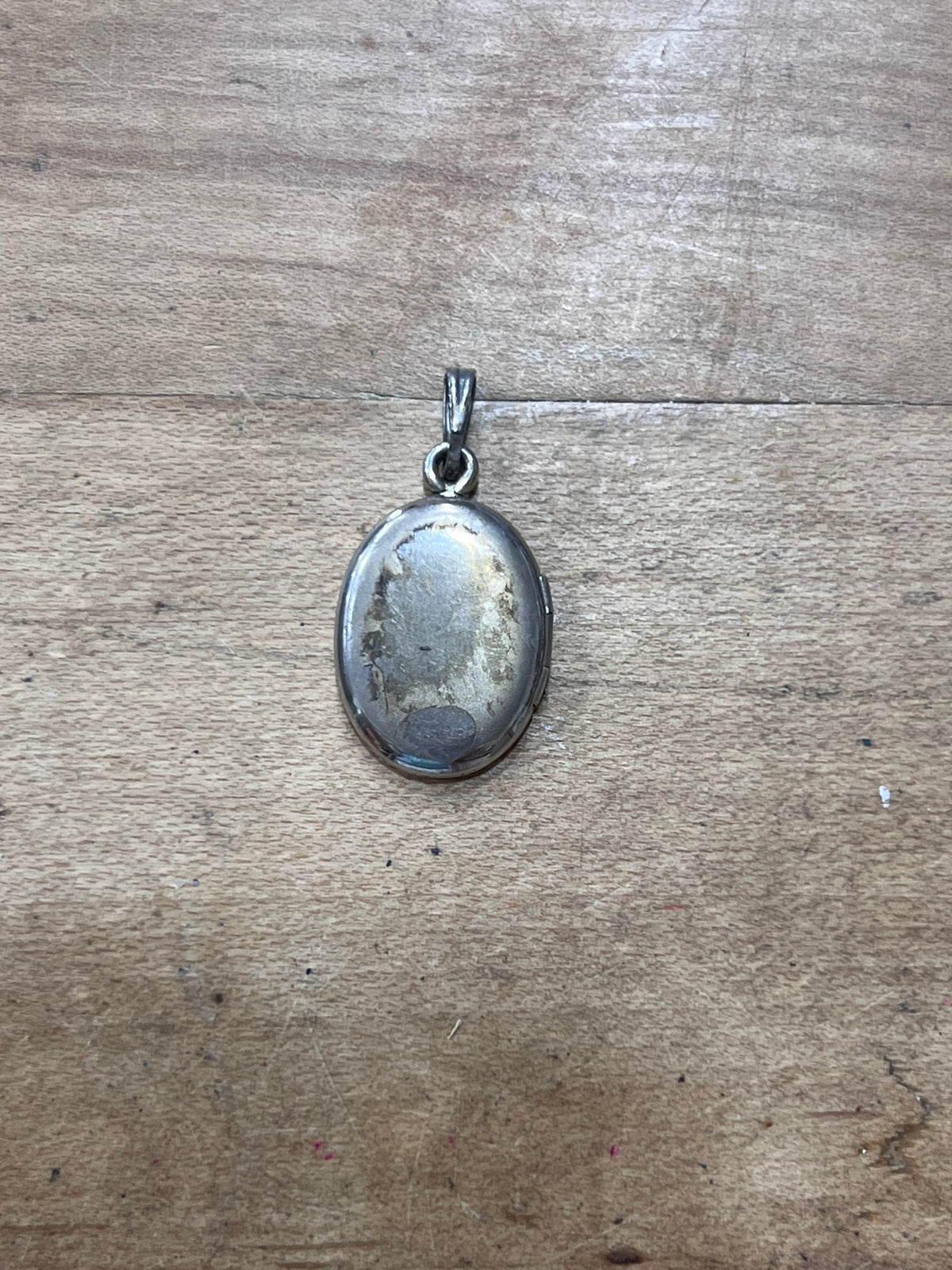 An oval silver pendant with a weathered, patinated surface, resting on a light brown wooden surface.