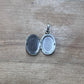 An open oval silver locket pendant with a bail, resting on a light brown wooden surface.