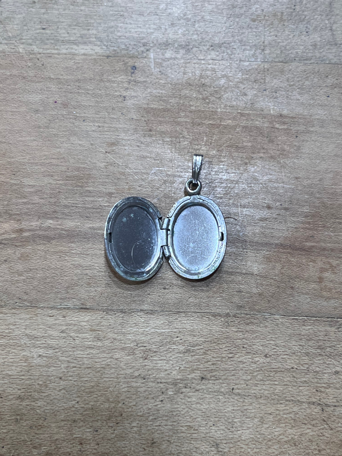 An open oval silver locket pendant with a bail, resting on a light brown wooden surface.
