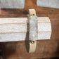 A beige leather wrap bracelet with a rectangular clear crystal cluster centerpiece and small gold-tone metal accents, displayed on a rustic wooden stand.