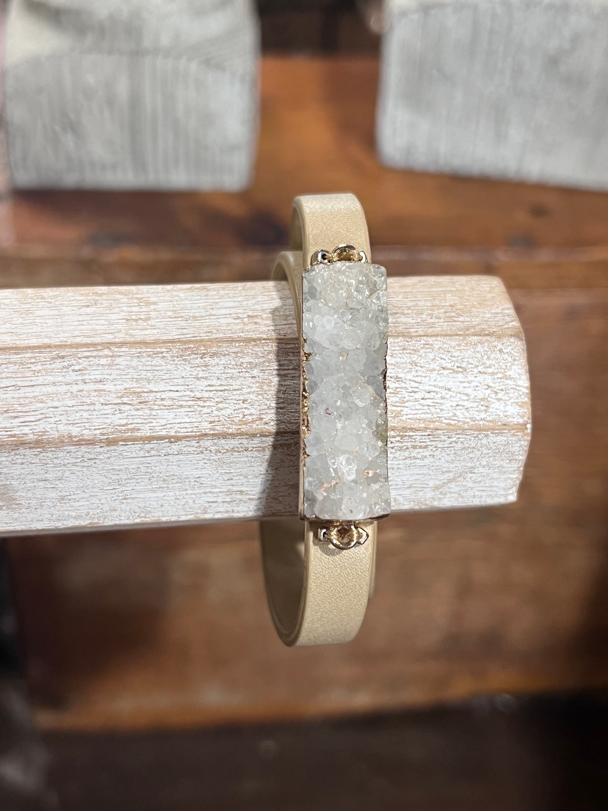 A beige leather wrap bracelet with a rectangular clear crystal cluster centerpiece and small gold-tone metal accents, displayed on a rustic wooden stand.