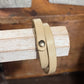 A beige leather wrap bracelet with a small gold-toned snap button, displayed on a light wood block stand.