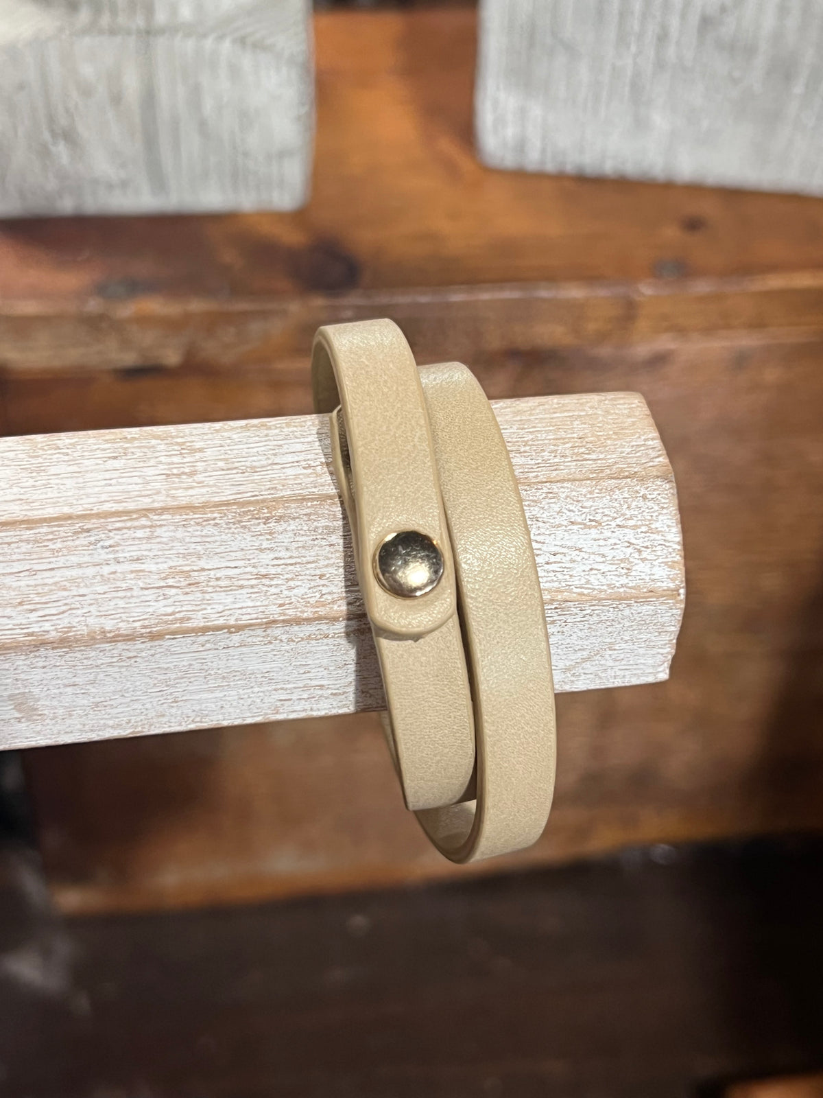 A beige leather wrap bracelet with a small gold-toned snap button, displayed on a light wood block stand.