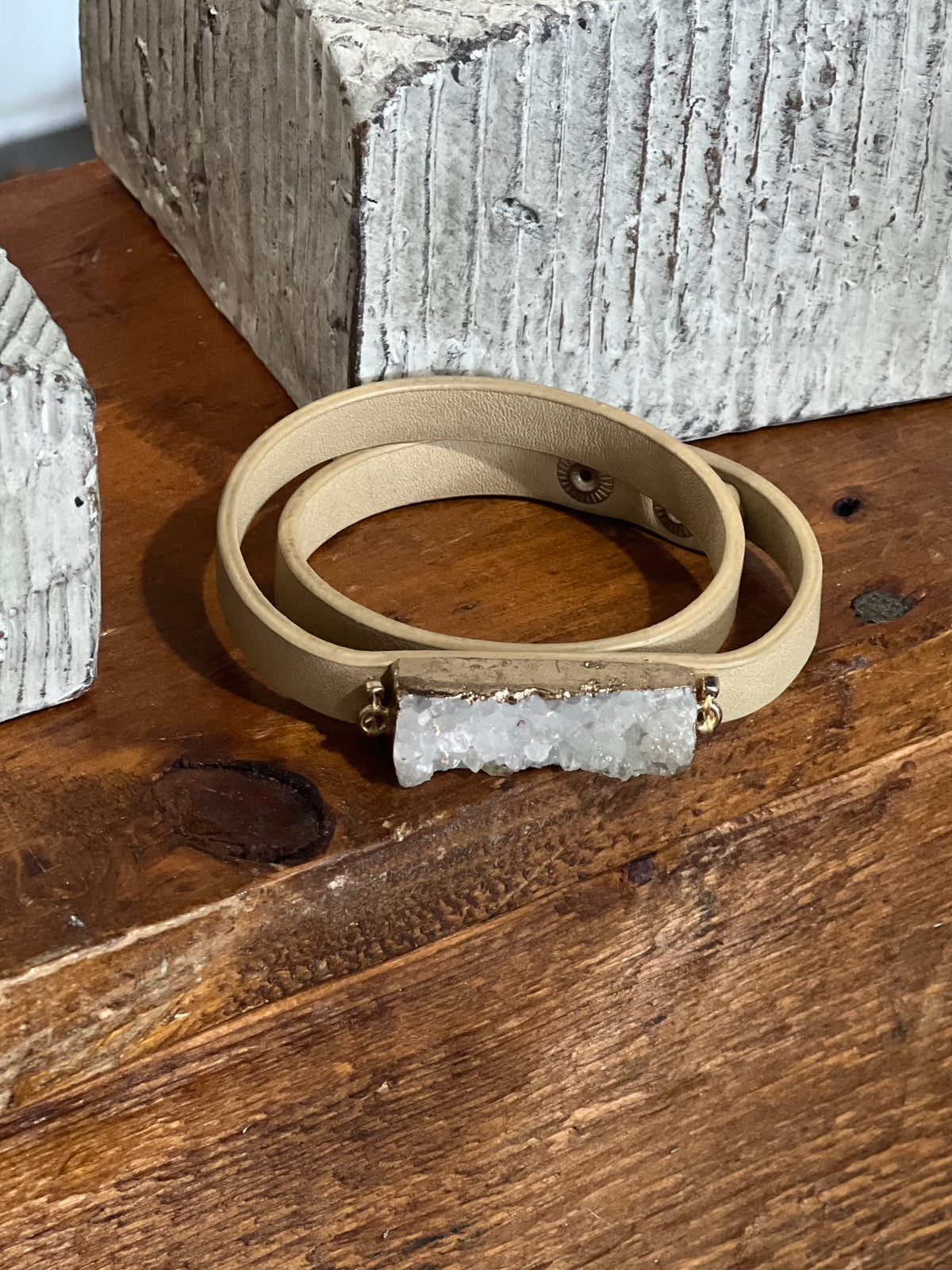 A beige leather wrap bracelet with a rectangular white crystal cluster centerpiece and small gold-tone metal accents, displayed on a rustic wooden surface.