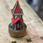 A miniature decorative house figurine with red roofs, gold trim, and a brown base, photographed on a wooden workbench.