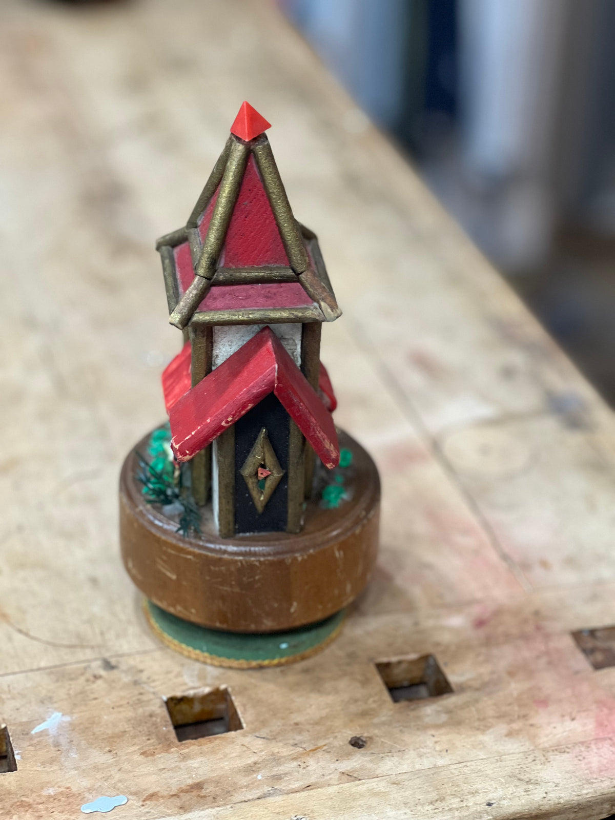 A miniature decorative house figurine with red roofs, gold trim, and a brown base, photographed on a wooden workbench.