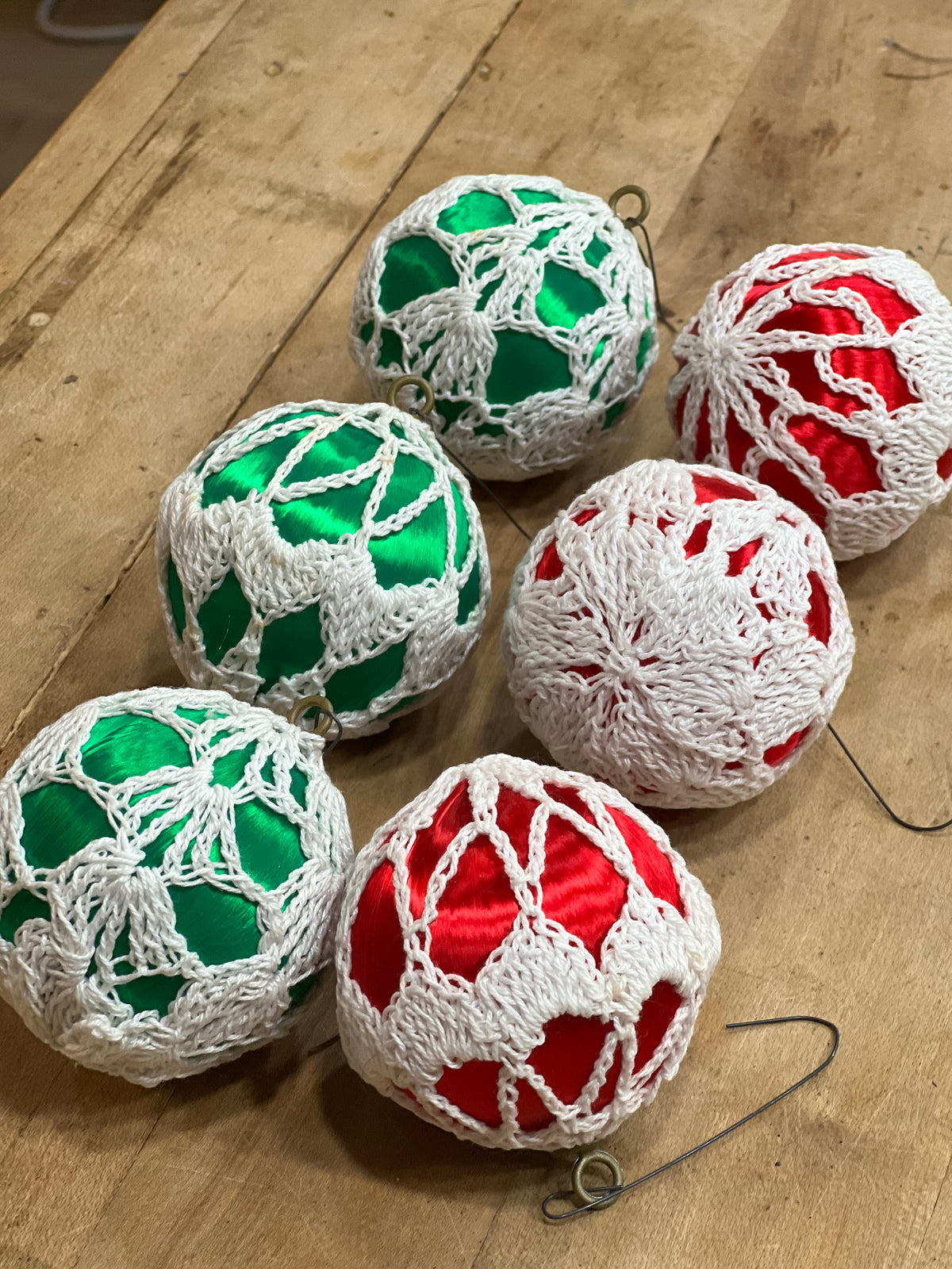 6 decorative Christmas ornaments wrapped in white crochet lace over shiny green and red satin balls, arranged on a wooden tabletop.