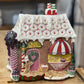 A decorative gingerbread-style house ornament with a candy shop theme, featuring a white icing-draped roof, pink door, striped awning, and cupcake details, displayed on a wooden surface.