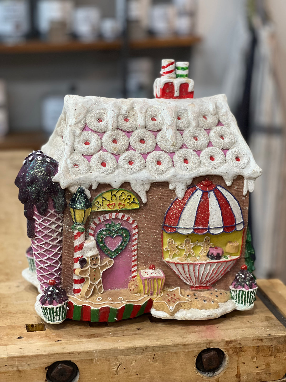 A decorative gingerbread-style house ornament with a candy shop theme, featuring a white icing-draped roof, pink door, striped awning, and cupcake details, displayed on a wooden surface.