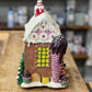 A gingerbread-style house ornament or decorative figurine with a snow-covered roof, pink icing details, a yellow window, and a large ice cream cone motif on the right side, displayed on a wooden surface with a blurred shop background.