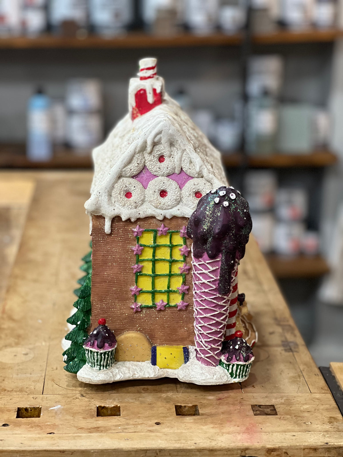 A gingerbread-style house ornament or decorative figurine with a snow-covered roof, pink icing details, a yellow window, and a large ice cream cone motif on the right side, displayed on a wooden surface with a blurred shop background.