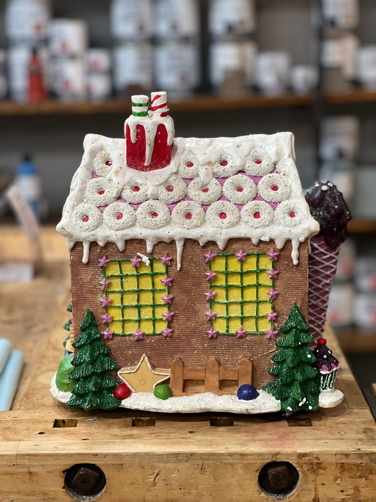 A gingerbread-style house ornament or decorative figurine with a white icing roof, green window panes, small green trees, a star, and candy-like details, displayed on a wooden surface with a blurred shop background.