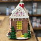 A small gingerbread-style Christmas house ornament or miniature house displayed on a wooden workbench, with shelves of colorful items blurred in the background.