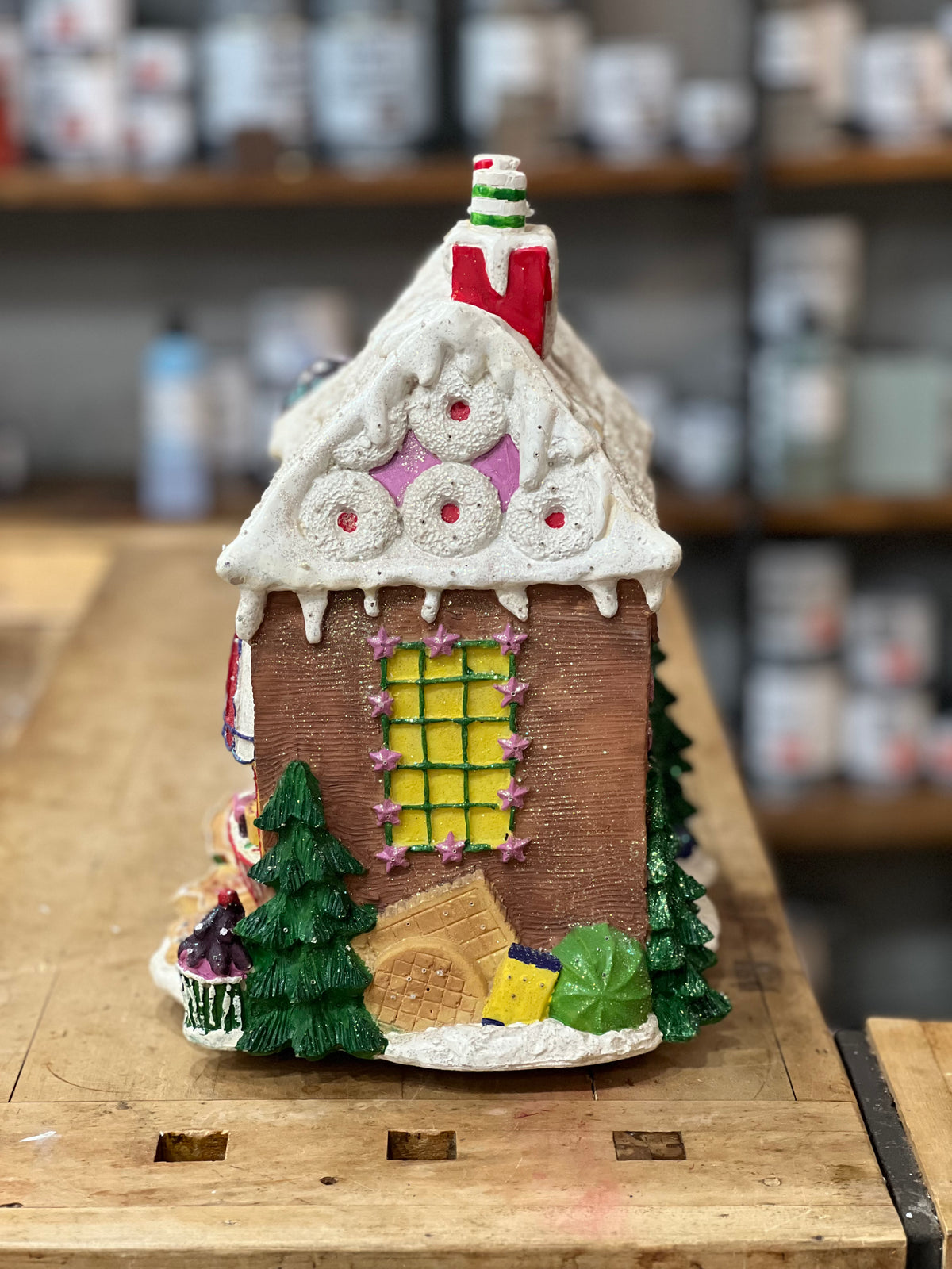 A small gingerbread-style Christmas house ornament or miniature house displayed on a wooden workbench, with shelves of colorful items blurred in the background.