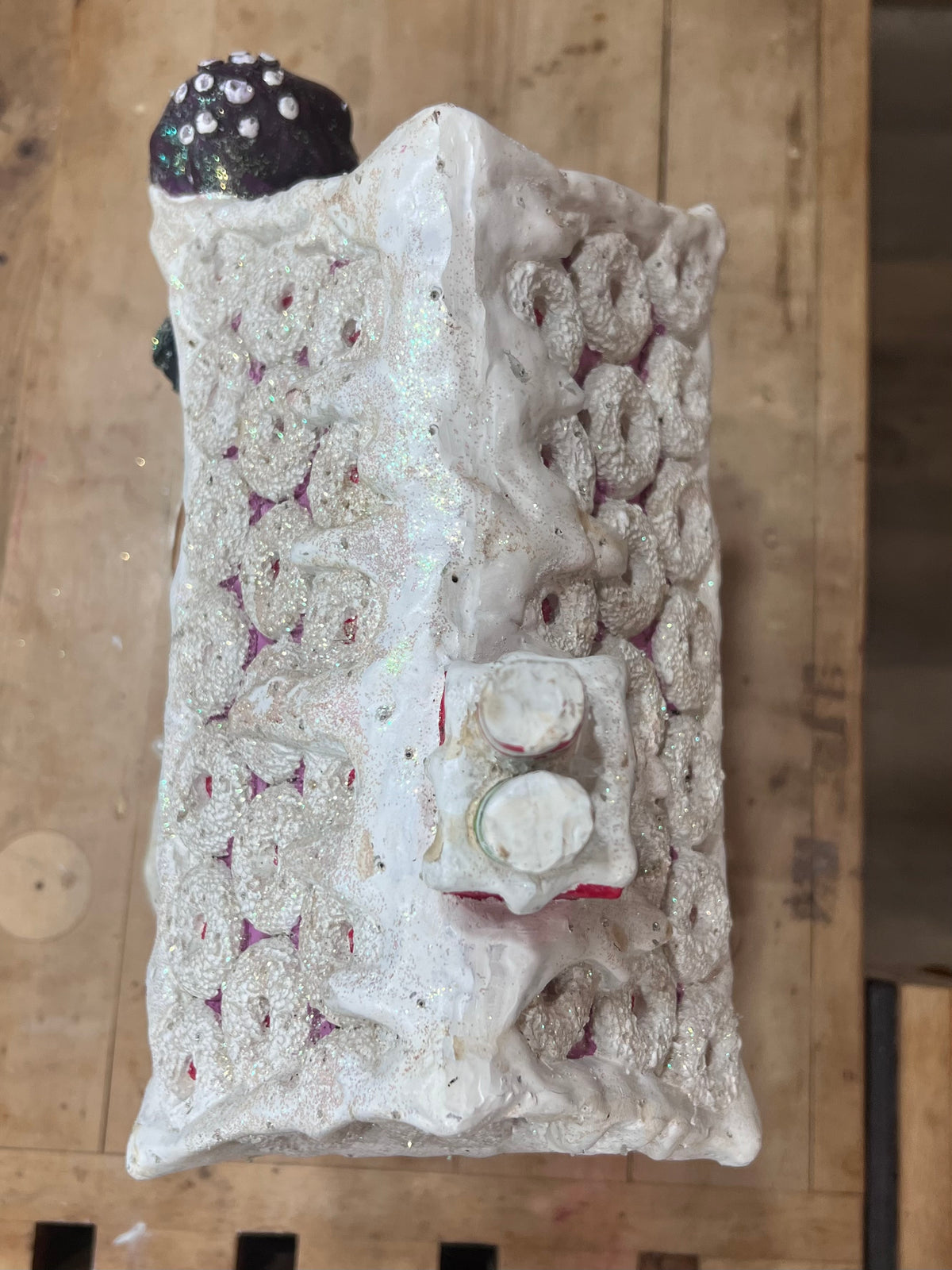 A textured, handcrafted rectangular object with a white and silver glittery surface, featuring a small dark top section with white dots, photographed on a wooden surface.