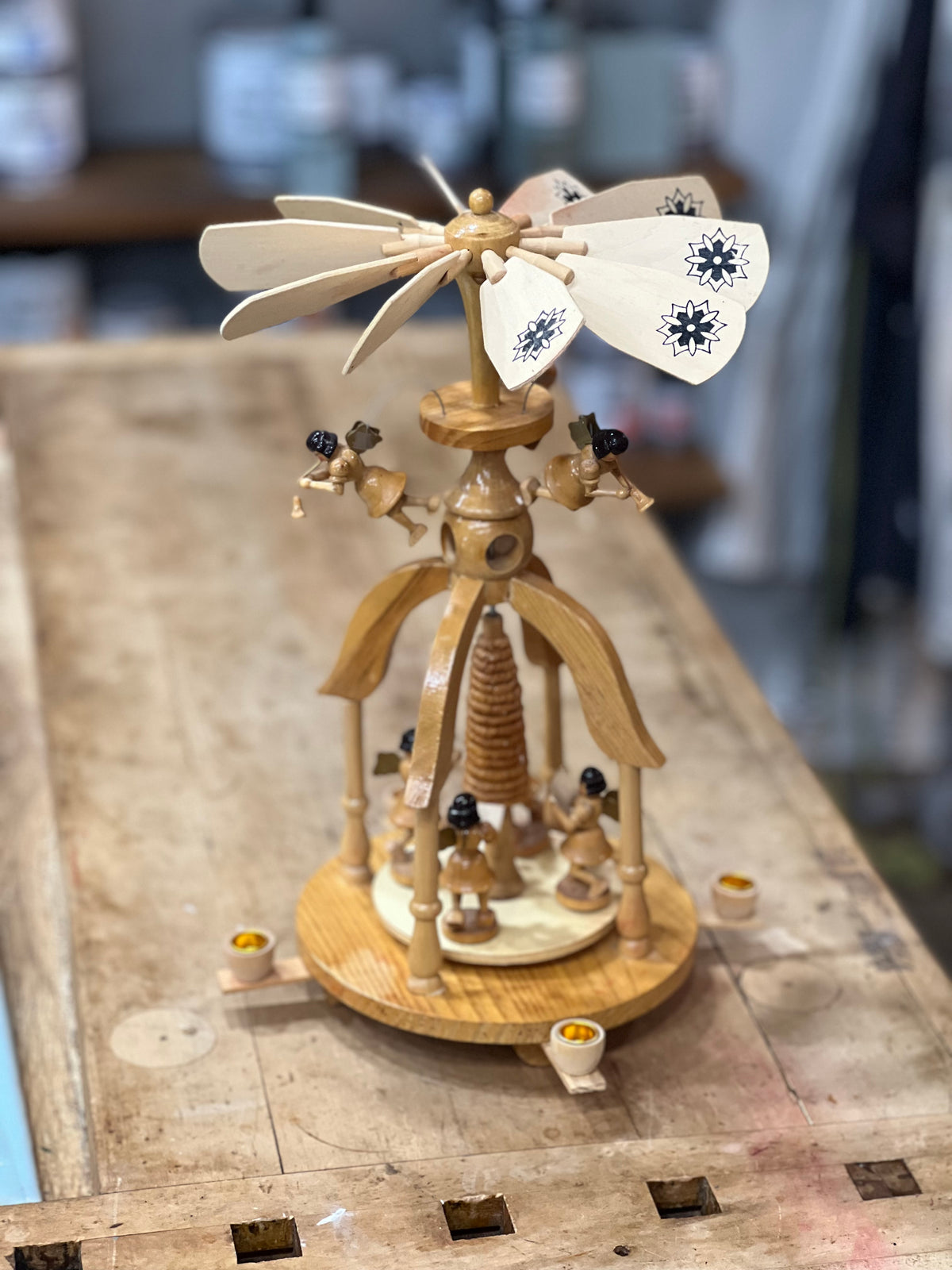 A handcrafted wooden spinning top with decorative painted blades and small figurines, displayed on a rustic wooden workbench.