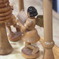 A close-up of a small handcrafted wooden figurine of an angel reading, standing on a round wooden base next to carved wooden tree trunks.