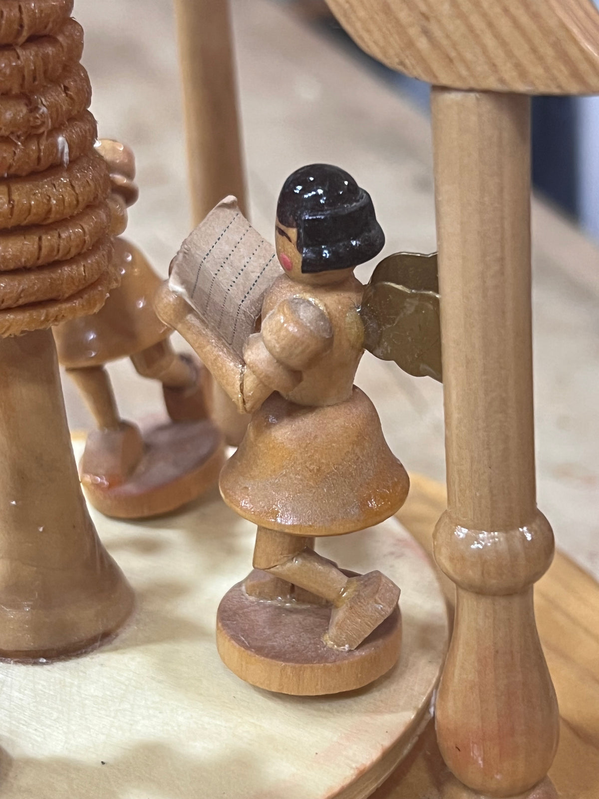 A close-up of a small handcrafted wooden figurine of an angel reading, standing on a round wooden base next to carved wooden tree trunks.