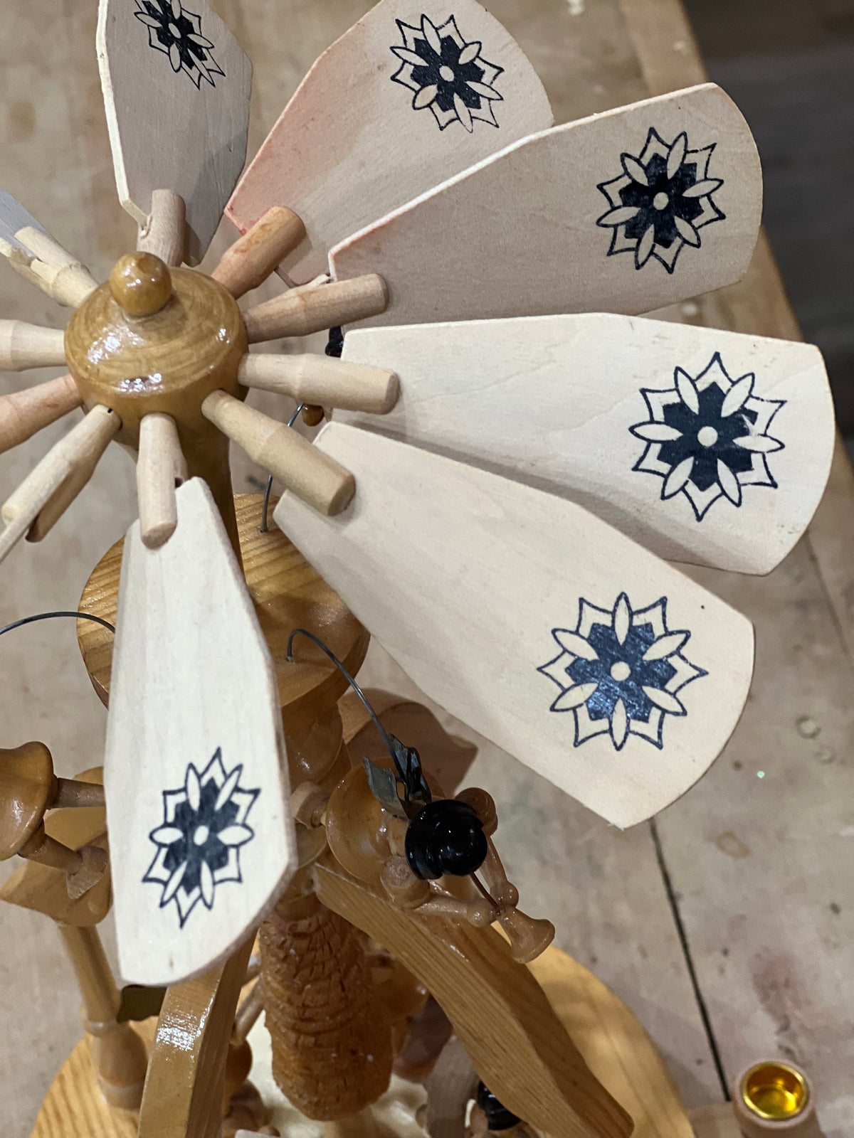 A close-up of a handcrafted wooden spinning wheel with light-colored wooden blades decorated with black floral star patterns, set against a workshop background.
