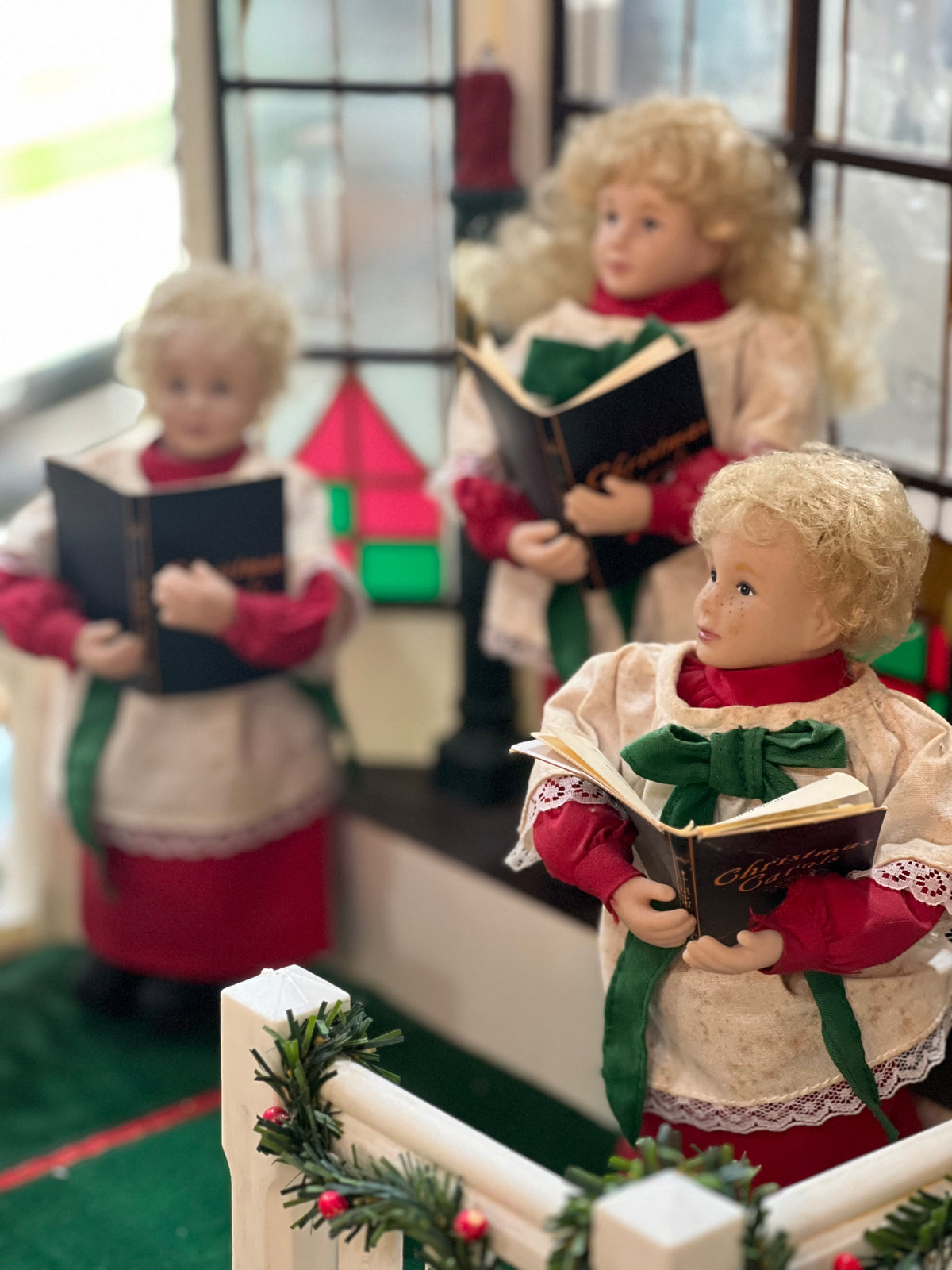 Three Christmas angel dolls dressed in red and cream outfits with green bows, each holding an open book, arranged indoors near a window and holiday garland.
