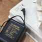 A black AC adapter power supply with gold text, shown next to a white plastic case on a wooden surface.