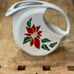 A white ceramic pitcher with a red poinsettia and green holly design, resting on a rustic wooden workbench.