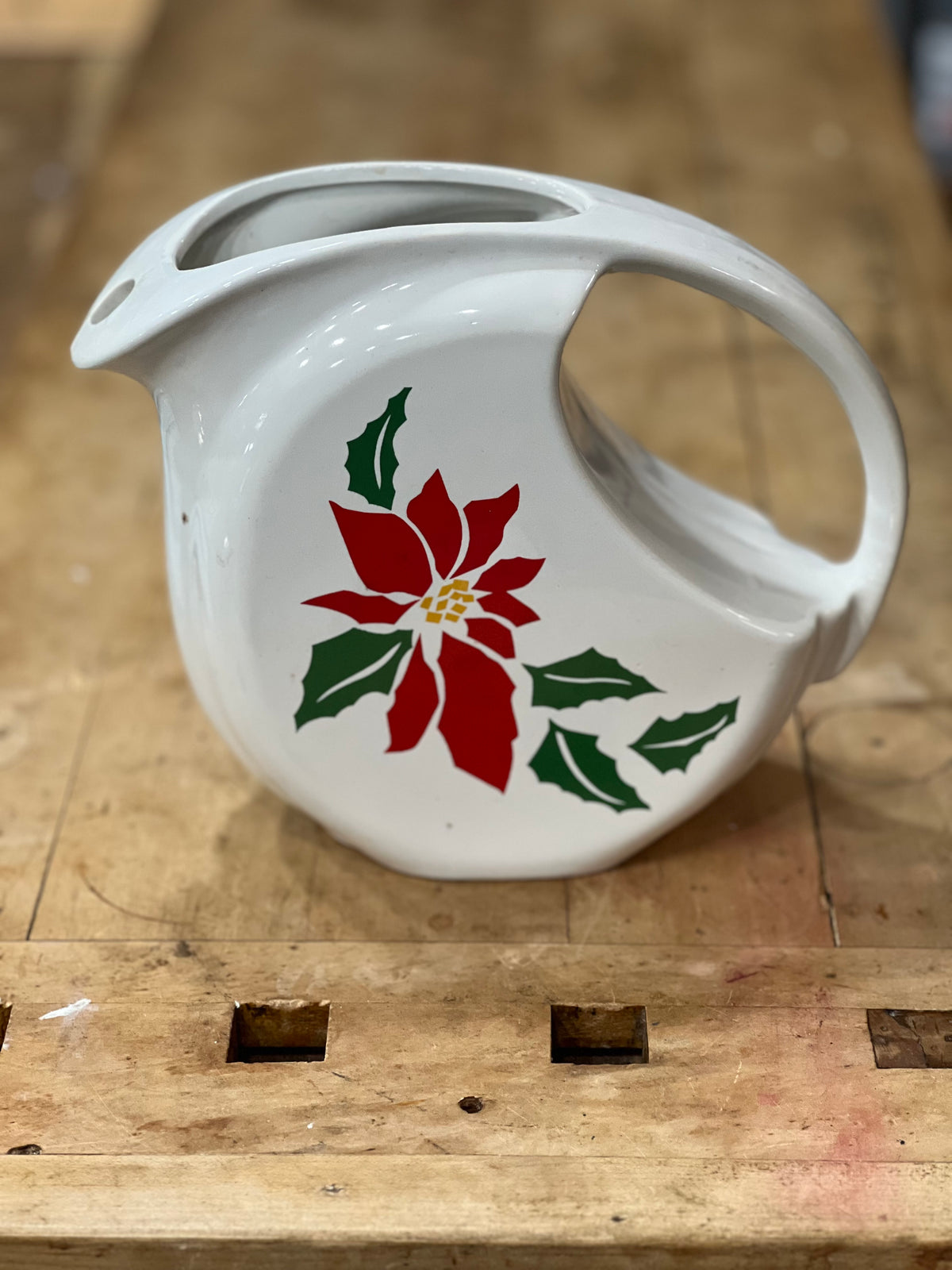 A white ceramic pitcher with a red poinsettia and green holly design, resting on a rustic wooden workbench.
