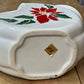 A white ceramic Christmas cookie dish with a red poinsettia and green holly design on the top, shown from the bottom to reveal a gold-colored brand stamp. The dish is placed on a wooden tabletop.