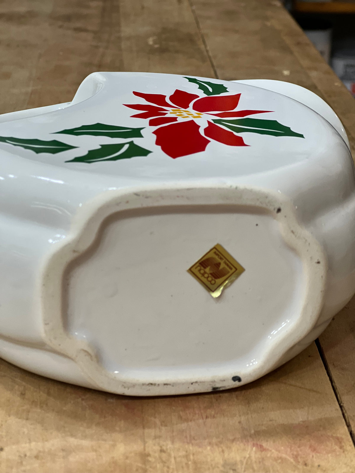 A white ceramic Christmas cookie dish with a red poinsettia and green holly design on the top, shown from the bottom to reveal a gold-colored brand stamp. The dish is placed on a wooden tabletop.