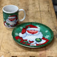 A Christmas-themed ceramic mug and matching plate set on a rustic wooden workbench. The designs feature a festive illustration of Santa Claus with snowflakes and ornaments.