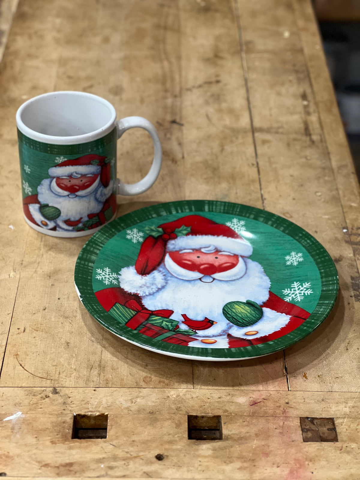 A Christmas-themed ceramic mug and matching plate set on a rustic wooden workbench. The designs feature a festive illustration of Santa Claus with snowflakes and ornaments.