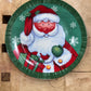 A round decorative Christmas plate featuring a painted Santa Claus with a red hat and coat, white beard, red gift boxes, and snowflakes on a green background, placed on a rustic wooden surface.