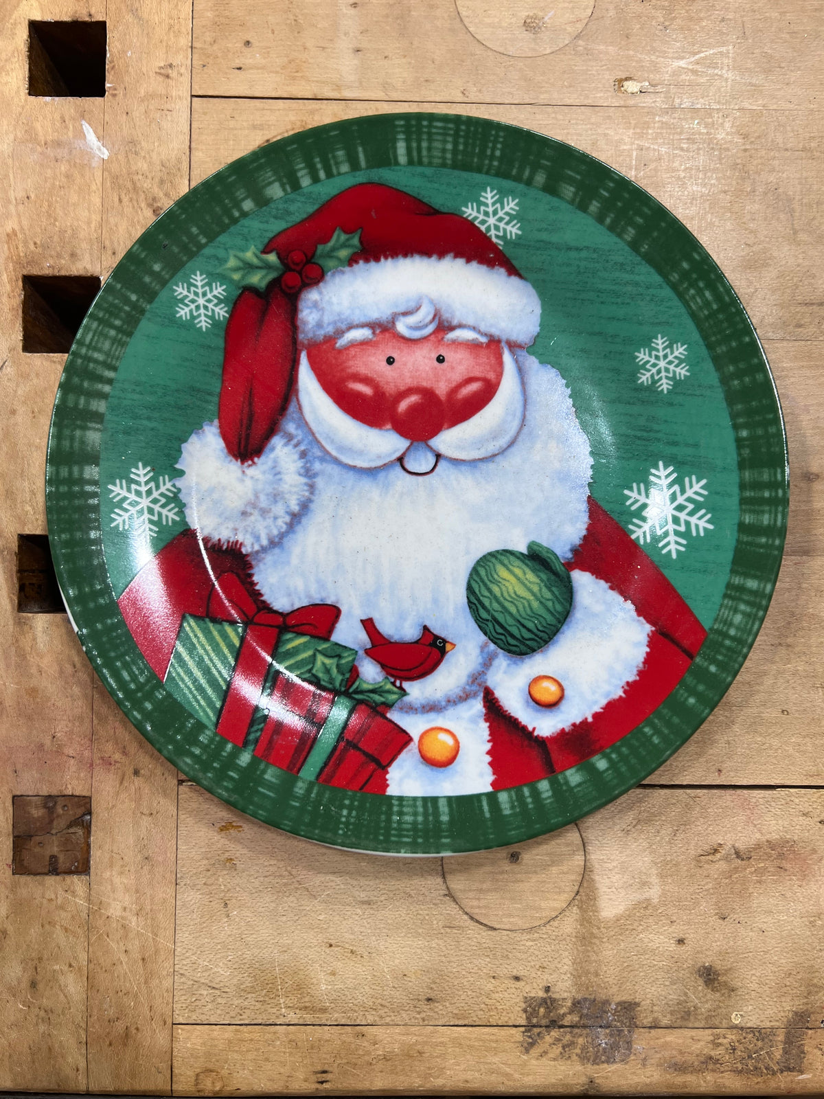 A round decorative Christmas plate featuring a painted Santa Claus with a red hat and coat, white beard, red gift boxes, and snowflakes on a green background, placed on a rustic wooden surface.