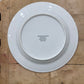 A white ceramic dinner plate shown from the back, resting on a rustic wooden workbench. The inside of the plate features printed black text with the brand 'Dorchester & McKinney Sons' and location details.