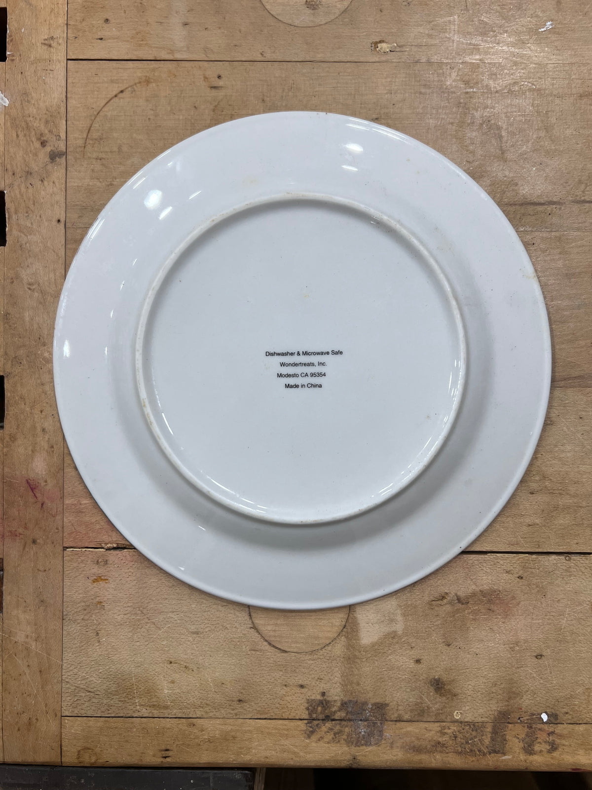 A white ceramic dinner plate shown from the back, resting on a rustic wooden workbench. The inside of the plate features printed black text with the brand 'Dorchester & McKinney Sons' and location details.