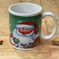 A white ceramic mug featuring a festive Christmas design of Santa Claus on a green background, placed on a wooden workbench.