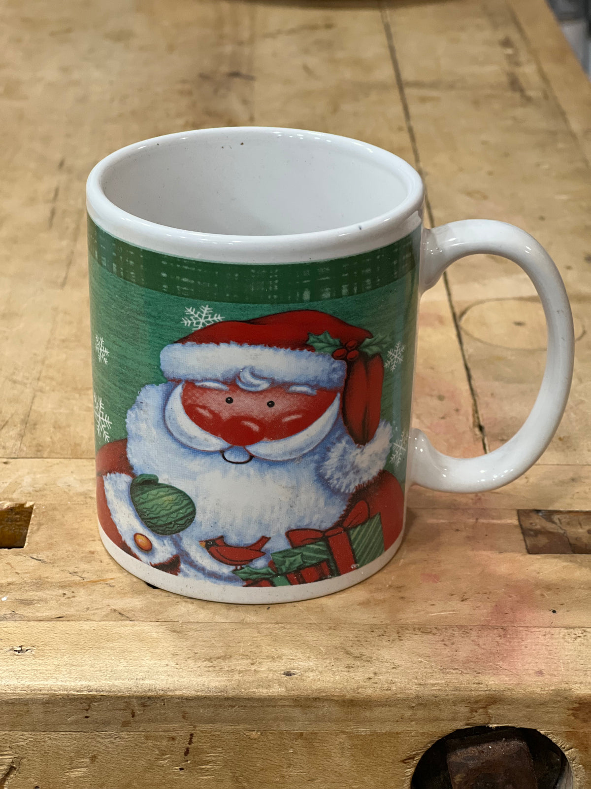 A white ceramic mug featuring a festive Christmas design of Santa Claus on a green background, placed on a wooden workbench.