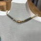 A beaded necklace displayed on a white mannequin bust, featuring dark green cylindrical beads, small gold spacer beads, and cream-colored end beads.