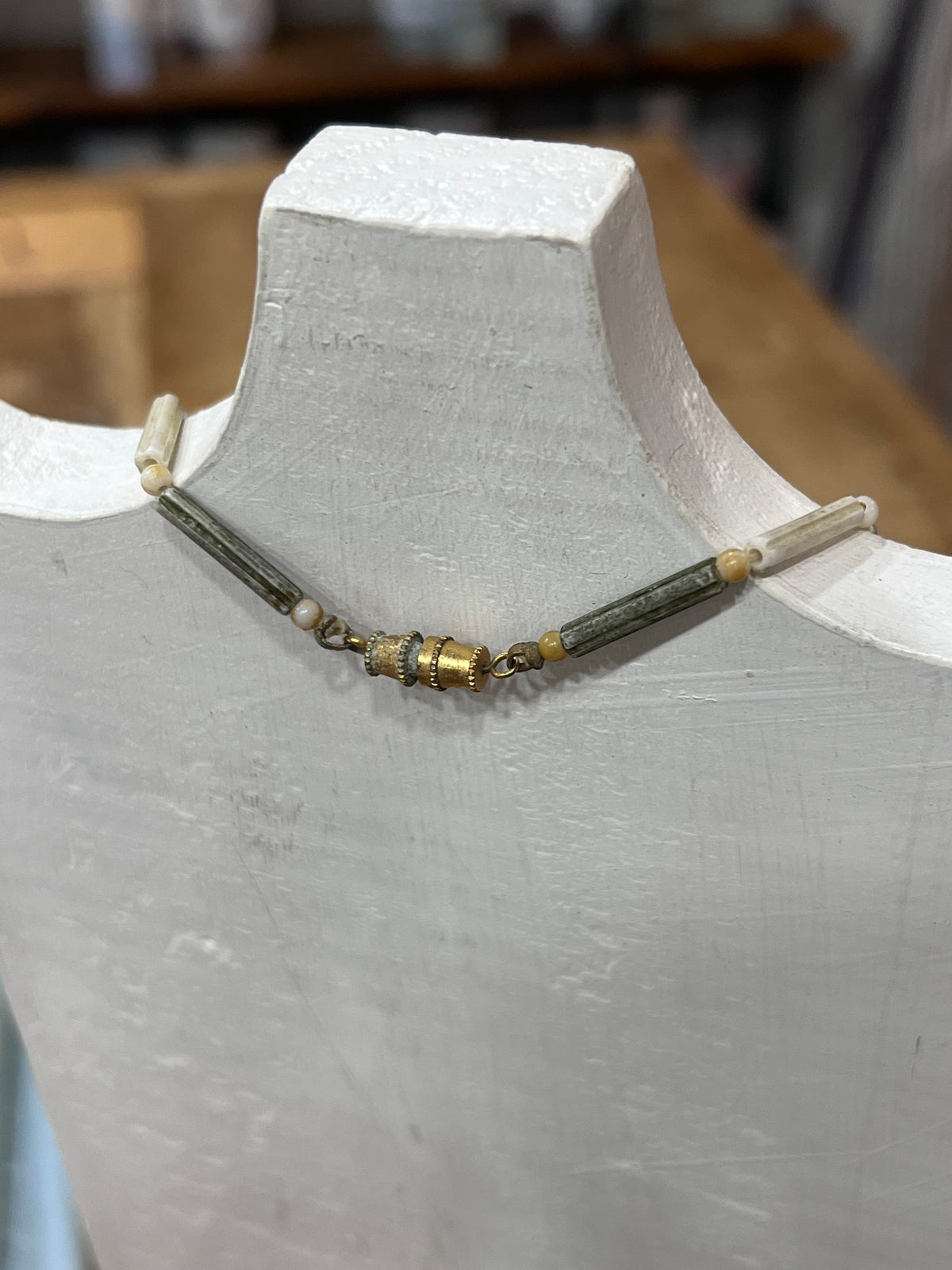 A beaded necklace displayed on a white mannequin bust, featuring dark green cylindrical beads, small gold spacer beads, and cream-colored end beads.