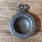 A vintage metal paint strainer with a hanging loop, featuring engraved text including 'HUGH REILLY COMPANY' and 'GLASS PAINTS - NATIONAL 1703', resting on a rustic wooden surface.