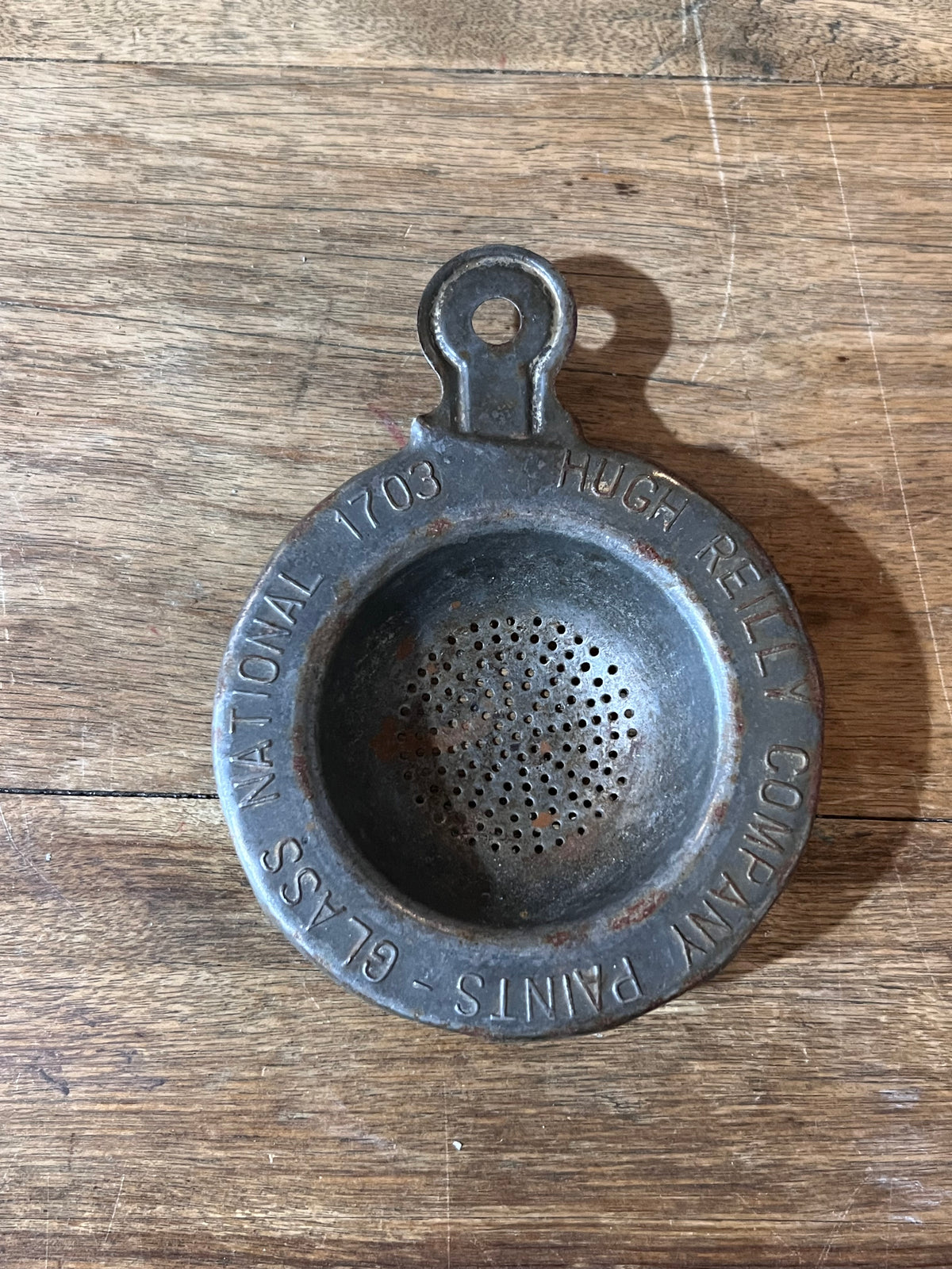 A vintage metal paint strainer with a hanging loop, featuring engraved text including 'HUGH REILLY COMPANY' and 'GLASS PAINTS - NATIONAL 1703', resting on a rustic wooden surface.
