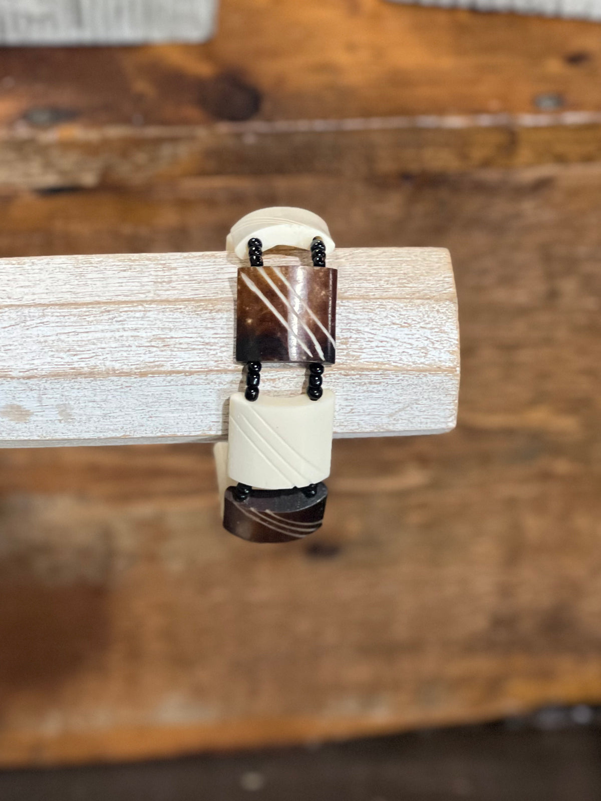 A close-up of a ring with a cream-colored textured band and dark brown marbled stone sections, displayed on a light-colored wooden block over a rustic wooden background.