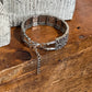 A silver-toned metal cuff bracelet with intricate cutout patterns and a dangling chain clasp, displayed on a rustic wooden surface next to a textured white stone prop.